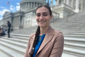 A woman in a brown suit jacket and blue shirt poses for a picture on the steps of the U.S. Capitol Building.