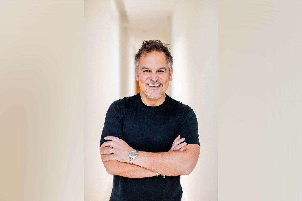 A man poses for a photo while wearing a black t-shirt. He arms are crossed.